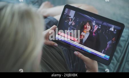 Tablet-Bildschirm zeigt die Wiedergabe von Nachrichten mit asiatischen Korrespondenten. Die kaukasische Frau sieht Nachrichtenberichte über US-Wahlen mit einem Tablet-Computer, während sie auf dem Sofa liegt. Nahaufnahme. Stockfoto