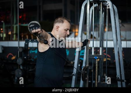 Ein Mann in einem schwarzen Tank-Top arbeitet mit einem Kabel-Gerät in einem Fitnessstudio. Stockfoto