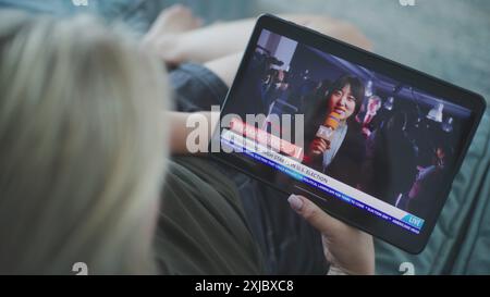 Tablet-Bildschirm zeigt die Wiedergabe von Nachrichten mit asiatischen Korrespondenten. Die kaukasische Frau sieht Nachrichtenberichte über US-Wahlen mit einem Tablet-Computer, während sie auf dem Sofa liegt. Nahaufnahme. Stockfoto