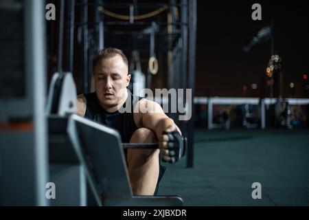 Ein Mann in einem schwarzen Tanktop trainiert nachts in einem Fitnessraum im Freien auf einem Rudergerät. Stockfoto