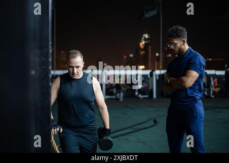 Ein Mann, der ein schwarzes Tanktop und eine schwarze Shorts trägt, hebt Gewichte, während ein Mann mit einem blauen Hemd zusieht. Stockfoto