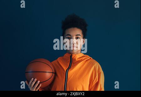 Porträt eines jungen Basketballspielers in oranger Sportkleidung, der im Studio posiert. Frau mit einem Basketball, der vor blauem Hintergrund steht. Porträt junger Menschen Stockfoto