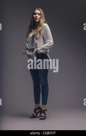 Selbstbewusste junge Frau in stilvollem Outfit posiert vor grauem Studiohintergrund und strahlt Eleganz und Trendlichkeit aus, mit Händen in den Taschen Stockfoto