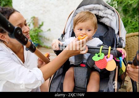 Mutter füttert ein Baby im Kinderwagen, draußen. Das Baby hält ein Stück Brot in der Hand, während farbenfrohe Spielzeuge in Fruchtform am Kinderwagen hängen. Ein warmes und liebevolles Hotel Stockfoto