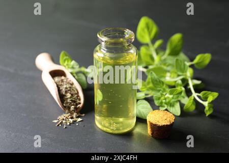 Ätherisches Öl in der Flasche, Löffel mit trockenem Kraut und Oregano-Zweigen auf dunkel strukturiertem Tisch, Nahaufnahme Stockfoto