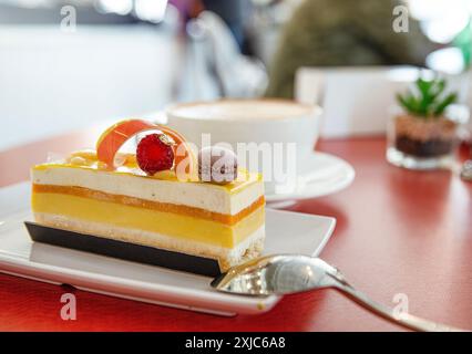 Ein Stück mehrschichtiges Dessert, garniert mit einer kleinen Frucht und Macaron, steht auf einem weißen Teller neben einem Löffel, mit einer Tasse Cappuccino im Hintergrund. Stockfoto