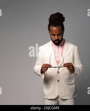 Der schicke Erwachsene latin-Mann mit Dreadlocks in Anzug und rosa Hemd sieht selbstbewusst aus, während er seine Brille hält Stockfoto