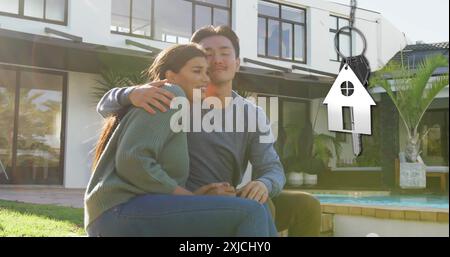 Bild des silbernen Hausschlüssels und Schlüsselanhängers über glücklichen, vielfältigen Paaren im Garten zu Hause Stockfoto