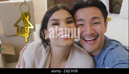 Bild von goldenem Hausschlüssel und Schlüsselanhänger über glückliches, vielfältiges Paar in einem neuen Zuhause Stockfoto