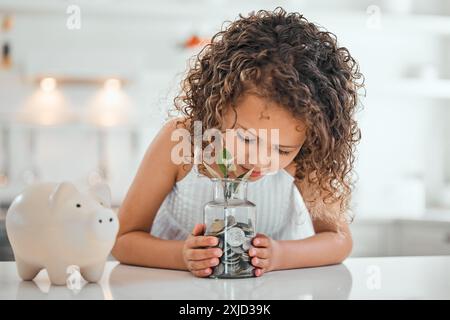 Kleines Mädchen, Geld und Münzen mit Glas oder Sparkasse für Ersparnisse, finanzielle Freiheit oder Wachstum zu Hause auf dem Tisch. Junge Frau, Kind oder Kind mit Stockfoto