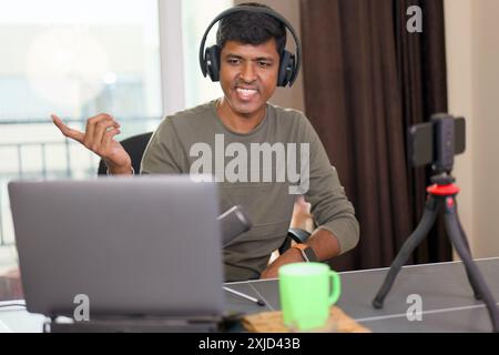 Ein lächelnder Mann bringt die Zuschauer während eines Livestreams zu Hause mit lebhaften Gesten in den Bann, indem er einen Laptop, Kopfhörer und ein Smartphone verwendet. Stockfoto