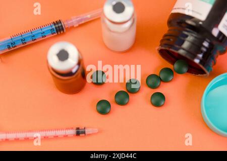 Grüne Tabletten, die aus einer Flasche neben Spritzen und Durchstechflaschen auf einem leuchtend orangefarbenen Hintergrund verschüttet werden. Stockfoto