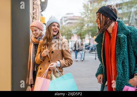 Fröhliche Gruppe von Frauen, die in urbaner Umgebung einkaufen. Freunde lachen und binden sich, während sie bunte Taschen tragen, die in stilvollen Herbstoutfits gekleidet sind Stockfoto