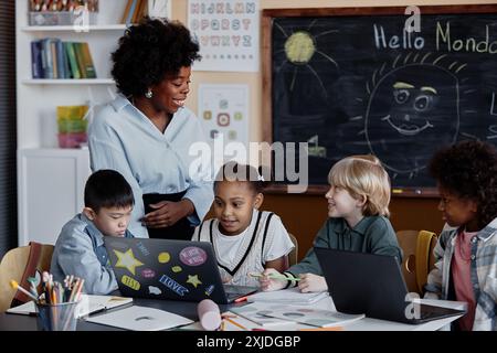 Empathische Lehrerin lächelt und beantwortet Kinderfragen während des Unterrichts in der Grundschule mit Laptops in verschiedenen Gruppen von Kindern, Kopierraum Stockfoto