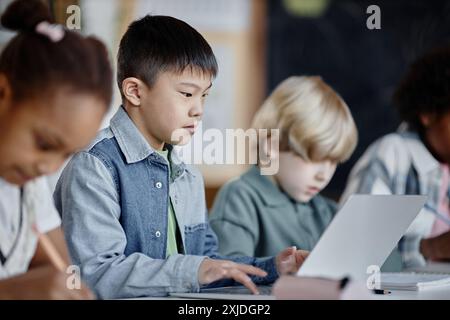 Seitliches Porträt eines asiatischen Jungen der Grundschule, der auf Laptop lernt, während er mit Klassenkameraden in Reihe sitzt Stockfoto