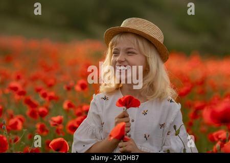 Ein junges blondes Mädchen steht auf einem Feld mit roten Mohnblumen und hält eine Blume. Sie lächelt und genießt die wunderschöne Landschaft Stockfoto