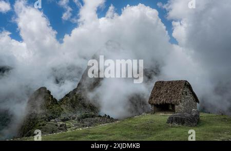 Ein malerischer Blick auf das historische Machu Picchu mit einer traditionellen Steinhütte und Wolken, die an einem sonnigen Tag die Berge bedecken, Peru Stockfoto