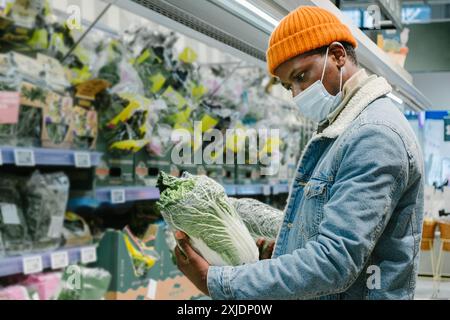 Afroamerikaner mit Maske und orangefarbener Beanie, kaufte frisches Gemüse in einem Supermarkt, prüfte verpacktes Gemüse. Stockfoto