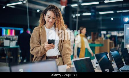 Porträt einer glücklichen hispanischen Frau, die ein Smartphone benutzt und mit Freunden in einer Online Social Media App SMS sendet. Kunden kaufen einen Laptop in einem Home Electronics Store Stockfoto