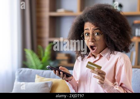 Schockierte Frau mit Brille, die Kreditkarte und Smartphone in der Hand hält, während sie im Wohnzimmer sitzt. Konzept unerwarteter Ausgaben, Online-Shopping, Finanzen, Banking, Überraschung, und Technologie. Stockfoto