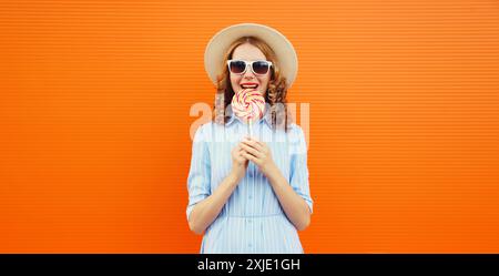 Sommerporträt einer glücklichen jungen Frau, die einen bunten Lutscher hält, ein fröhliches Mädchen im Touristenhut, das auf einem hellorangefarbenen Hintergrund posiert Stockfoto