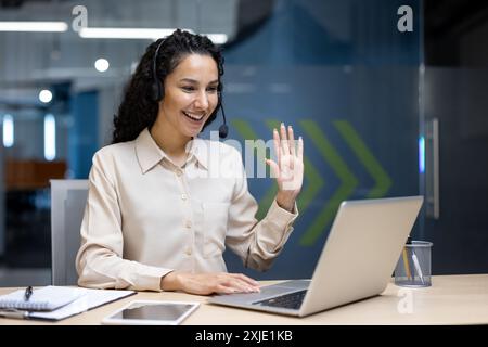 Lächelnde Frau mit Headset, die professionelle Videoanrufe in moderner Büroumgebung führt. Teilnahme an virtuellen Meetings, Arbeitsumgebung mit Laptop, Notebook und Stift am Schreibtisch. Stockfoto