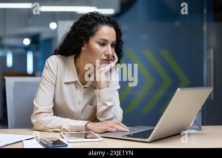 Junge professionelle Frau, die sich im modernen Büro auf Laptop konzentriert. Arbeitsumgebung mit elektronischen Geräten und Papierkram. Konzept von Geschäft, Technologie und Produktivität. Stockfoto