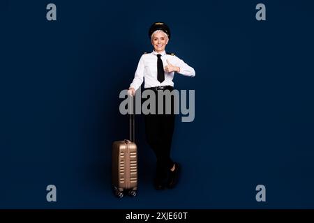 Foto in voller Größe von hübschen pensionierten weiblichen professionellen Flugzeugpiloten Koffer Daumen hoch tragen Pilotenuniform isoliert auf dunkelblauem Hintergrund Stockfoto