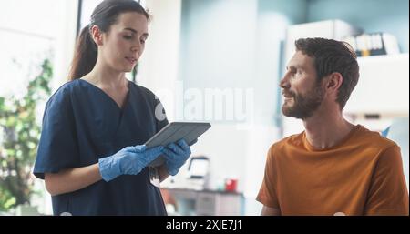 Kaukasischer Mann, Der Auf Dem Stuhl Im Hellen Öffentlichen Krankenhaus Sitzt Und Weibliche Krankenschwester Mit Tablet-Computer Hört. Professionelle Frau, Die Mögliche Nebenwirkungen Nach Der Medizininjektion Erklärt. Stockfoto