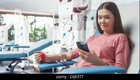 Kaukasierin Spendet Blut Für Menschen In Not. Spenderin Drückt Den Herzförmigen Roten Ball, Um Blut Zu Pumpen Und Online Mit Dem Smartphone Zu Chatten. Spende Für Frauen Mit Schwangerschaftskomplikationen. Stockfoto