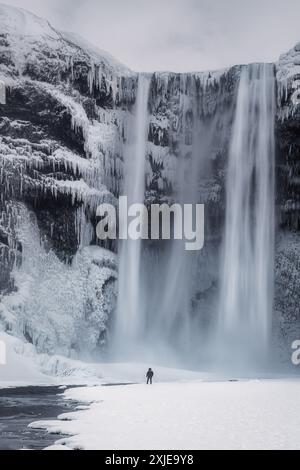 Ein einsamer Wanderer vor dem massiven majestätischen Wasserfall Skogafoss im Süden Islands, an einem Wintermorgen, mit Schnee und Eis in der Schlucht, Reiseziel Stockfoto