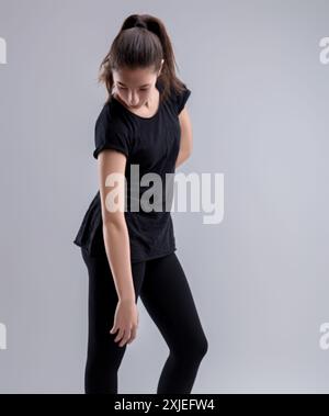 Selbstbewusste junge Frau in schwarzem T-Shirt und Leggings posiert anmutig vor grauem Hintergrund und verkörpert modernen Stil und gesunde Weiblichkeit Stockfoto