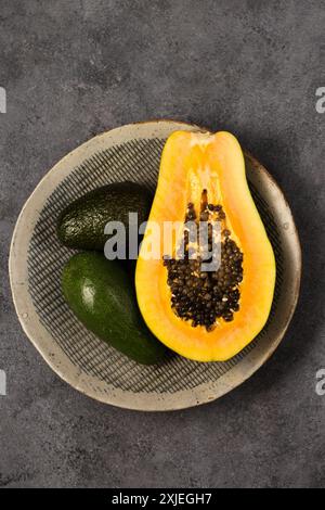 Zwei Avocados und eine halbe Papaya auf einer grauen Tonplatte von Hand auf grauem Hintergrund, Blick von oben Stockfoto