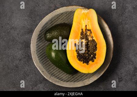 Zwei Avocados und eine halbe Papaya auf einer grauen Tonplatte von Hand auf grauem Hintergrund, Blick von oben Stockfoto