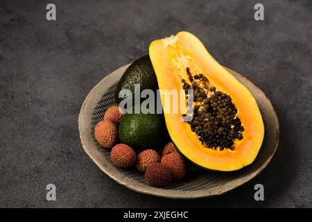 Zwei Avocados, eine halbe Papaya und Litschi-Beeren auf einer grauen Tonplatte von Hand auf grauem Hintergrund Stockfoto