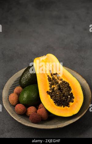 Zwei Avocados, eine halbe Papaya und Litschi-Beeren auf einer grauen Tonplatte von Hand auf grauem Hintergrund Stockfoto