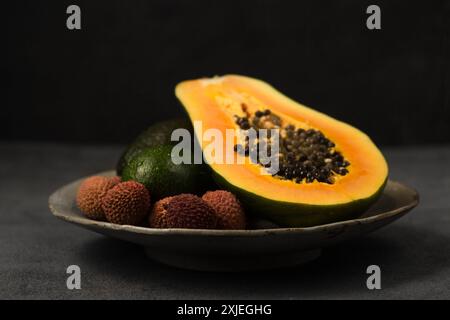 Zwei Avocados, eine halbe Papaya und Litschi-Beeren auf einer grauen Tonplatte von Hand auf grauem Hintergrund Stockfoto