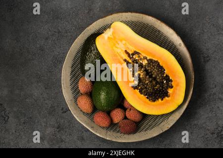 Zwei Avocados, eine halbe Papaya und Litschi-Beeren auf einer grauen Tonplatte von Hand auf grauem Hintergrund, Draufsicht Stockfoto