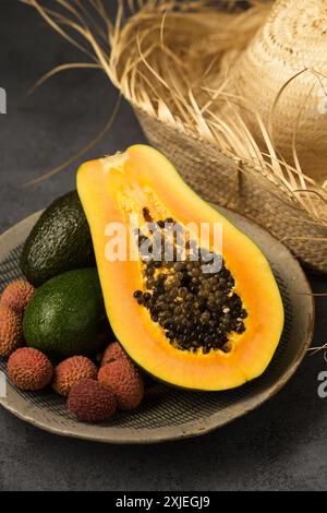 Zwei Avocados, eine halbe Papaya und Litschi-Beeren auf einer grauen Tonplatte, die von Hand in der Nähe des Strohhutes hergestellt wurde Stockfoto