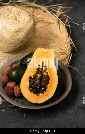 Zwei Avocados, eine halbe Papaya und Litschi-Beeren auf einer grauen Tonplatte, die von Hand in der Nähe des Strohhutes hergestellt wurde Stockfoto