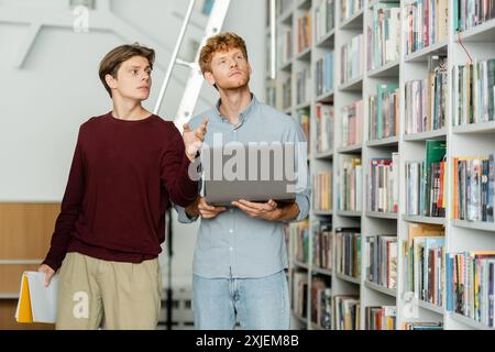 Zwei Männer, die in Laptopforschung vertieft sind, inmitten von Büchern in einer Bibliothek. Stockfoto