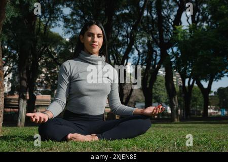 Junge Lateinfrau sitzt in Lotusblume Position im Park, sie wird nach dem Büro ausgeruht, verbindet sich mit der Natur, sieht glücklich in die Kamera, Kopierraum Stockfoto