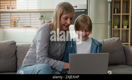 Lächelnde kaukasische Großmutter ältere 60er Jahre Frau hilft zu lernen glücklicher kleiner Enkel Kind Junge benutze Laptop zusammen Couch. Reife weibliche Seniorenfamilie Stockfoto