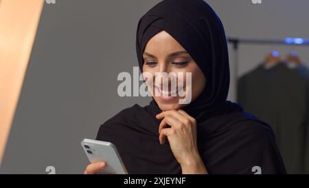 Glückliche islamische Frau Blick Bildschirm von Handy-Gerät fröhliche muslimische Mädchen in Hijab Lachen Wischen Feed soziale Netzwerk im Studio Arabische Frau genießen Stockfoto