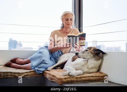 Seniorin, liest Buch und entspannt mit Hund auf Sofa zum Lernen, Wissen oder gemütlich zusammen im Wohnzimmer. Tier, Geschichte und älterer Mensch Stockfoto