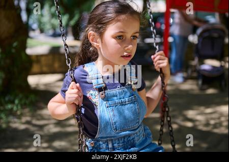 Ein junges Mädchen mit blauem Overall sitzt auf einer Schaukel und sieht an einem sonnigen Tag nachdenklich in einem Park aus. Hintergrund zeigt Grün und unscharfe Figuren. Stockfoto