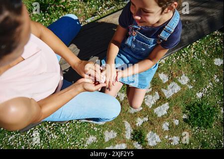 Blick aus einem Hochwinkel auf eine Mutter und Tochter, die Hände im Park halten. Die Tochter trägt Denim-Overalls und sitzt auf einer Holzbank. Stockfoto