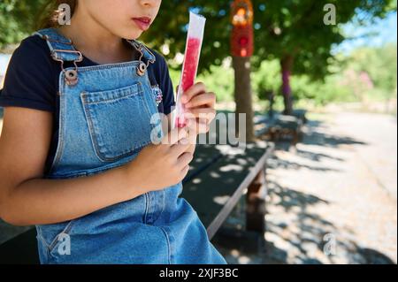 Ein junges Mädchen mit Denim-Overall und marineblauem Hemd genießt an einem sonnigen Tag in einem Park, umgeben von Natur und Bäumen, ein rotes Eis. Stockfoto