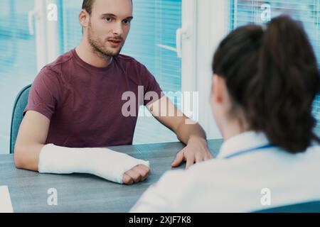 Mann mit gebrochenem Arm spricht mit einem Arzt im Krankenhaus. Sie diskutieren Behandlung und Pflege und betonen Kommunikation, Vertrauen und Empathie in der Gesundheitsversorgung Stockfoto