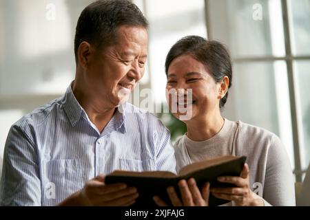 Glückliches älteres asiatisches Paar, das zu Hause auf der Familiencouch sitzt und zusammen Bibel liest Stockfoto